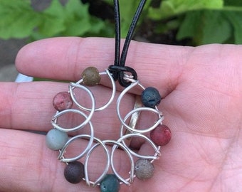 May include: A silver wire necklace with a circular pendant featuring a flower design. The pendant is adorned with eight small, round, multi-colored stones.