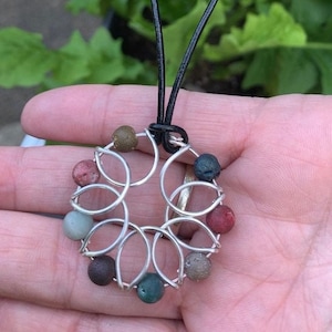 Puede incluir: Un collar de alambre plateado con un colgante circular con diseño de flor. El colgante está adornado con ocho pequeñas piedras redondas multicolores.