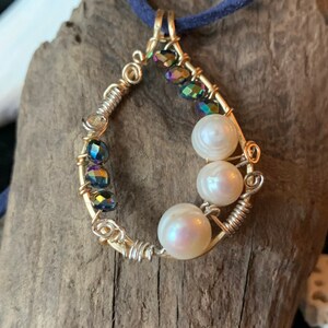 Könnte beinhalten: Ein goldfarbener, drahtgewickelter Anhänger mit schillernden Glasperlen und weißen Perlen. Der Anhänger hängt an einer dunkelblauen Schnur.