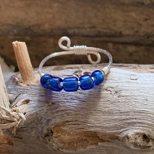 May include: A handmade silver wire ring featuring five vibrant blue glass beads. The ring's design includes a coiled wire section and decorative loops, set against a natural wood background. This piece of jewelry is a unique accessory.