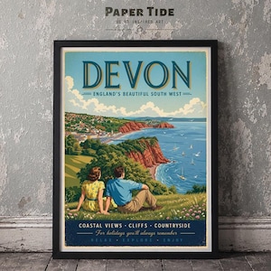 May include: A framed travel poster featuring a coastal landscape of Devon, England. The illustration shows two people sitting on a grassy hill overlooking red cliffs, a blue sea with sailboats, and a town. Text reads: DEVON, ENGLAND'S BEAUTIFUL SOUTH WEST.