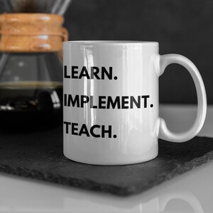 May include: White ceramic mug with black text that reads "LEARN. IMPLEMENT. TEACH."