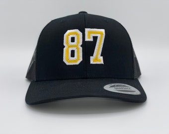 May include: Black trucker hat with a yellow and white embroidered number 87.