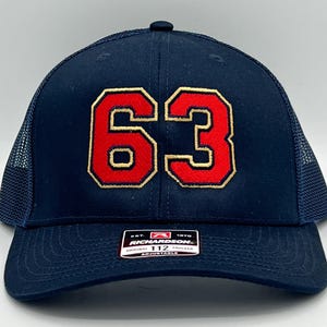 May include: Navy blue trucker hat with the number "63" in red with a gold outline. The hat has a mesh back and a curved brim. The Richardson brand label is visible on the front.