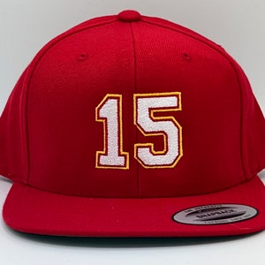 Puede incluir: Gorra de béisbol roja con un número 15 blanco bordado en la parte delantera. El número tiene un contorno amarillo. La gorra tiene un cierre de presión negro.