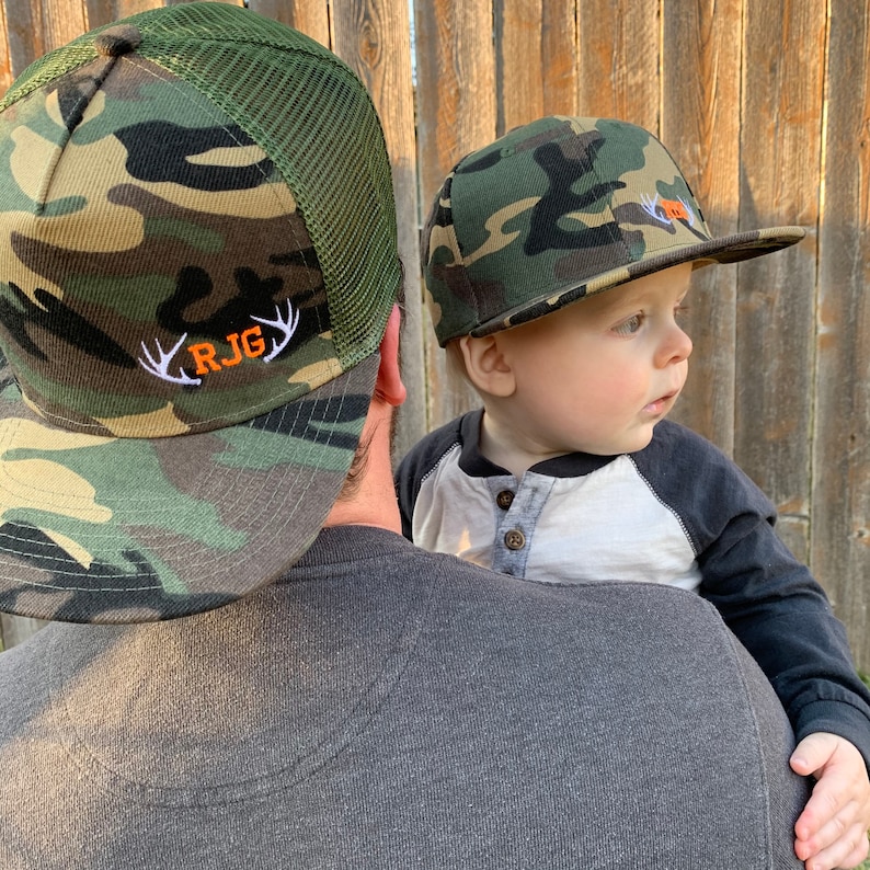 May include: A father and son wearing matching camouflage baseball caps with embroidered antlers and initials. The father's cap has the initials "RJG" and the son's cap has the initials "RJ".