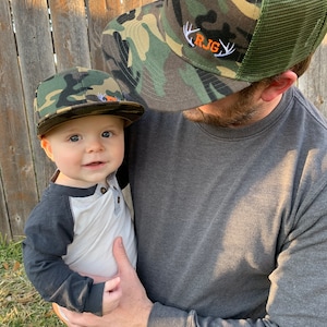 May include: A father and son wearing matching camouflage baseball caps. The father's cap has the initials "R.J.G." embroidered on the front with white antlers. The son's cap has a small orange patch on the front.