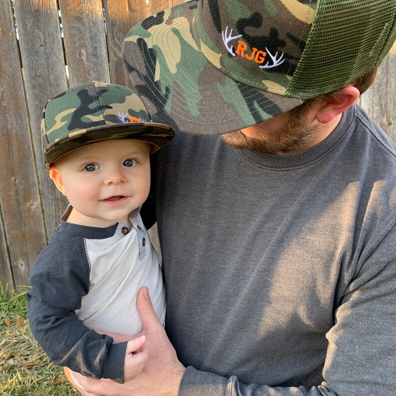 May include: A father and son wearing matching camouflage baseball caps. The father's cap has the initials "R.J.G." embroidered on the front with white antlers. The son's cap has a small orange patch on the front.