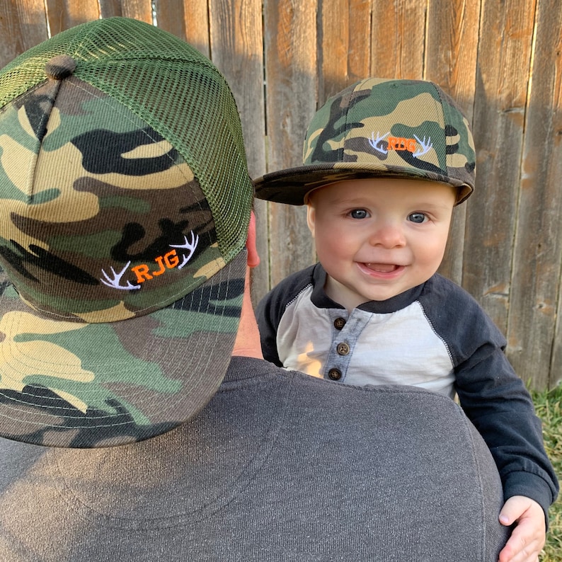May include: A father and son wearing matching camouflage baseball caps with embroidered antlers and initials. The father's cap says "RJG" in orange lettering. The son's cap has a small orange bear embroidered on it.