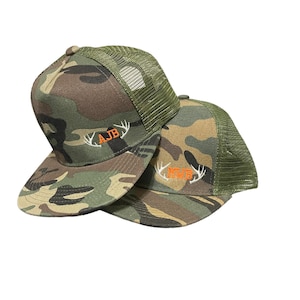 May include: Two camouflage trucker hats with mesh back panels. The top hat has the initials "AJB" embroidered in orange with antlers on either side. The bottom hat has the initials "MWB" embroidered in white with antlers on either side.