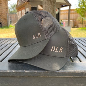 May include: Two dark green trucker hats with mesh panels. The front of each hat has a different embroidered name, "SLB" and "DLB".