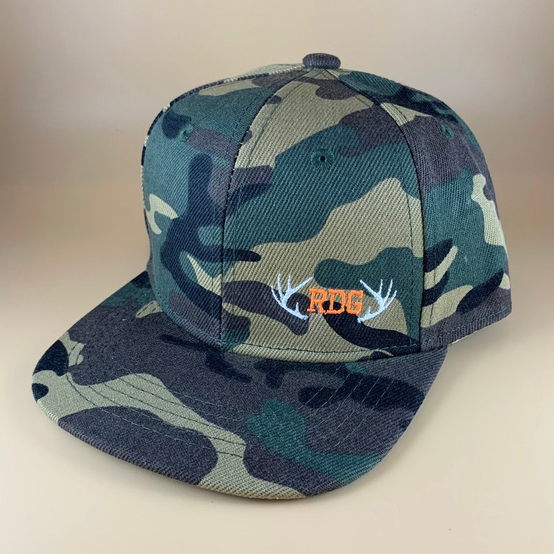 May include: Camouflage baseball cap with a white embroidered design of antlers and the letters "RBG" in orange.