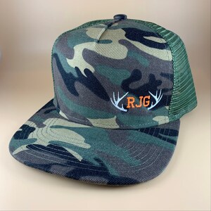 May include: Camouflage trucker hat with a white mesh back and an orange embroidered logo that says "RJG" with white antlers on either side.