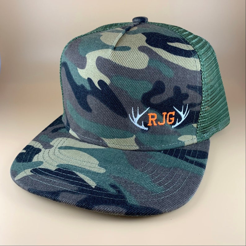 May include: Camouflage trucker hat with a white mesh back and an orange embroidered logo that says "RJG" with white antlers on either side.