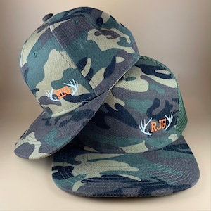 May include: Two camouflage baseball caps with embroidered antlers and initials. The top cap has the initials "RBG" and the bottom cap has the initials "RJG".