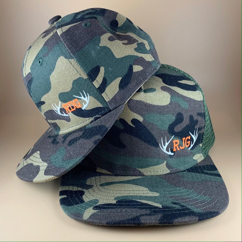 May include: Two camouflage baseball caps with embroidered antlers and initials. The top cap has the initials "RBG" and the bottom cap has the initials "RJG".