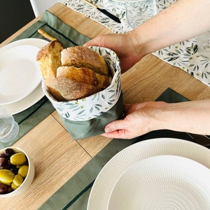 May include: A green fabric bread basket with a white and green olive branch pattern, holding sliced bread. The basket is on a wooden table with a green placemat and a white plate.