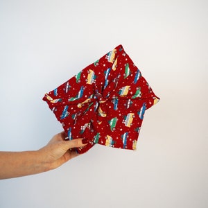 May include: A red fabric wrap with a pattern of colorful cars and white stars. The fabric is tied in a knot, creating a unique presentation. The wrap is held by a hand.