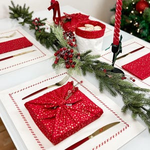 Op de afbeelding: Een feestelijke kersttafelsetting met een rood en goud sterrenpatroon verpakt cadeau, gouden bestek en rode servetten. De tafel is versierd met een groene guirlande, rode bessen en lange rode kaarsen. Een kerstboom is zichtbaar op de achtergrond.