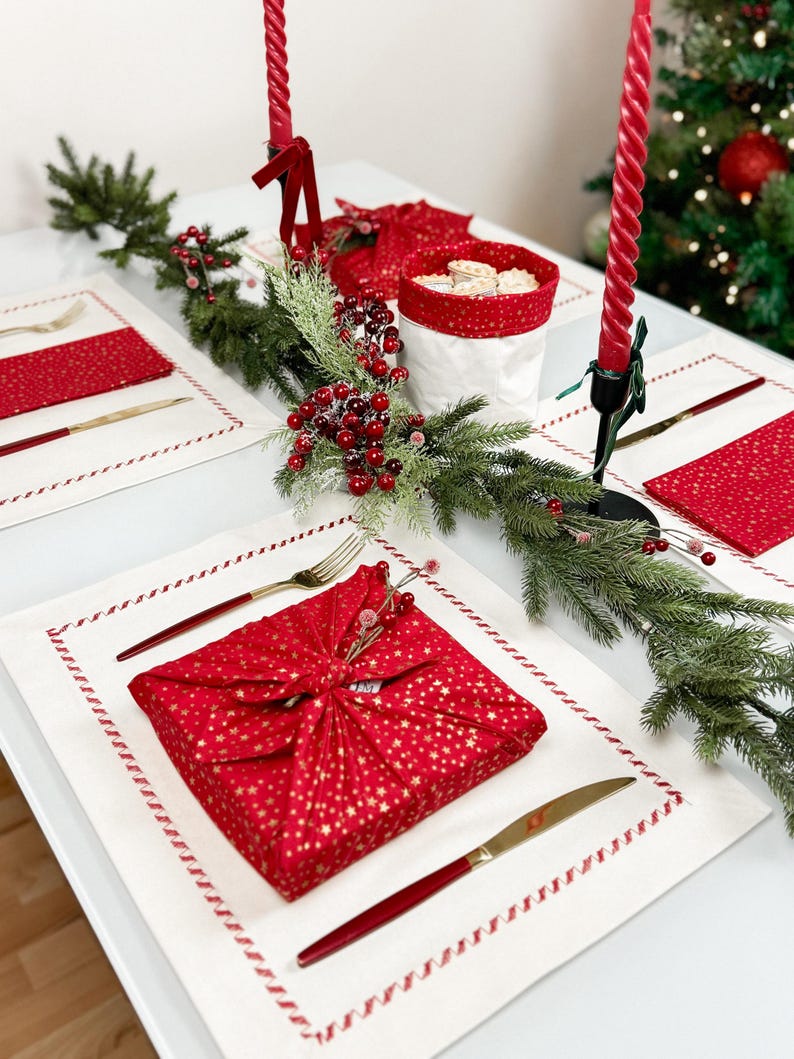 Op de afbeelding: Een feestelijke kersttafelsetting met een rood en goud sterrenpatroon verpakt cadeau, gouden bestek en rode servetten. De tafel is versierd met een groene guirlande, rode bessen en lange rode kaarsen. Een kerstboom is zichtbaar op de achtergrond.