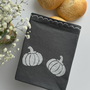 May include: A dark grey fabric basket featuring two white pumpkin designs. The top edge has a scalloped trim. Two golden-brown rolls are in the background, with a small basket of white flowers to the left.