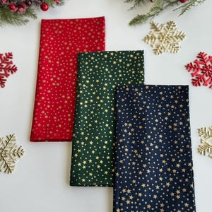 Op de afbeelding: Drie gevouwen stoffen servetten in rood, groen en marineblauw, elk versierd met een gouden sterrenpatroon. De servetten zijn gerangschikt op een wit oppervlak, versierd met feestelijke elementen zoals sneeuwvlokken en bessen takken.