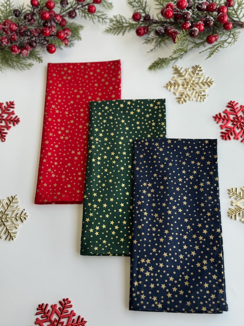 Op de afbeelding: Drie gevouwen stoffen servetten in rood, groen en marineblauw, elk versierd met een gouden sterrenpatroon. De servetten zijn gerangschikt op een wit oppervlak, versierd met feestelijke elementen zoals sneeuwvlokken en bessen takken.