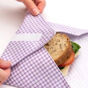 Puede incluir: Un envoltorio de tela a cuadros morado y blanco con un sándwich en su interior. El sándwich tiene un relleno de tomate, lechuga y queso.