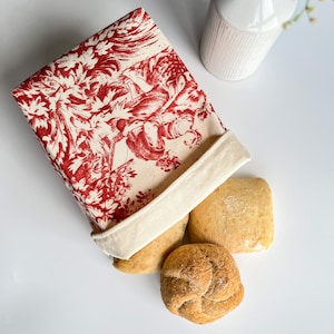 Puede incluir: Una bolsa de pan de tela con estampado rojo y blanco con tres panes dentro. La bolsa está hecha de algodón y tiene un cierre de cordón blanco.