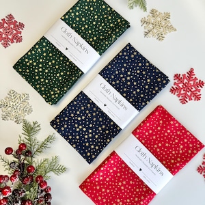 Op de afbeelding: Drie gevouwen stoffen servetten in groen, marineblauw en rood, elk versierd met gouden sterrenpatronen. Elke servet is verpakt met een witte band met de tekst "Cloth Napkins". Feestelijke kerstversieringen omringen de servetten.