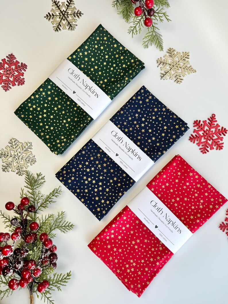 Op de afbeelding: Drie gevouwen stoffen servetten in groen, marineblauw en rood, elk versierd met gouden sterrenpatronen. Elke servet is verpakt met een witte band met de tekst "Cloth Napkins". Feestelijke kerstversieringen omringen de servetten.