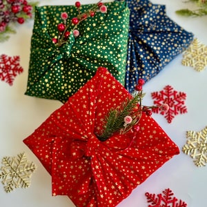 Op de afbeelding: Drie in stof verpakte cadeaus in groen, rood en marineblauw, elk versierd met gouden sterren. De groene en rode pakketten zijn versierd met bessen en groen. Gouden en rode sneeuwvlokornamenten zijn rond de cadeaus verspreid.