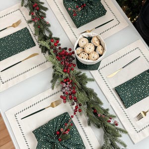Op de afbeelding: Een feestelijke tafelsetting met cr&egrave;mekleurige placemats, gouden vorken en messen en groene servetten met witte sterren. Een groen met stof verpakt cadeau en een mandje met mini-taarten zijn ook aanwezig, samen met een slinger van groen en rode bessen.