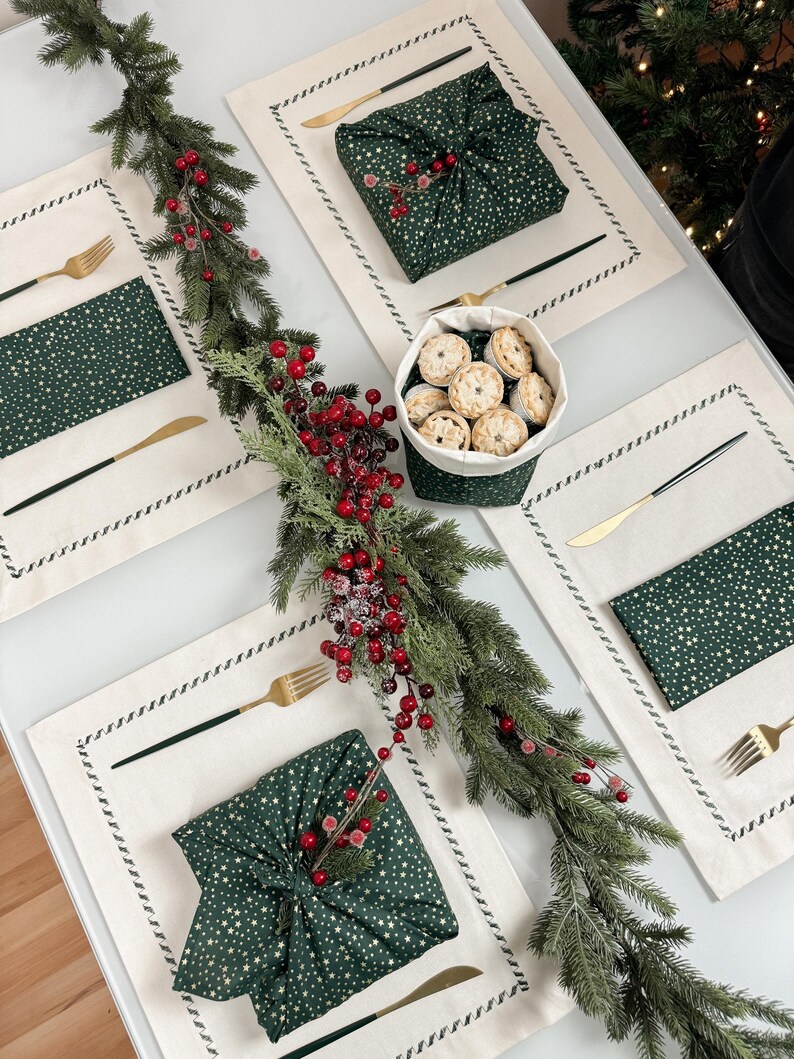 Op de afbeelding: Een feestelijke tafelsetting met cr&egrave;mekleurige placemats, gouden vorken en messen en groene servetten met witte sterren. Een groen met stof verpakt cadeau en een mandje met mini-taarten zijn ook aanwezig, samen met een slinger van groen en rode bessen.