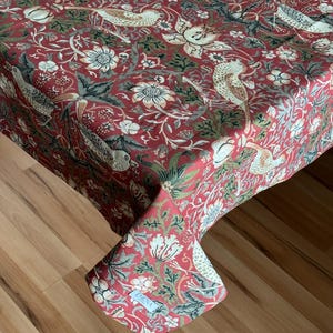 Może przedstawiać: Czerwony obrus z bogatym wzorem ptaków, kwiatów i liści. Wzór zawiera białe, zielone i szare akcenty na czerwonym tle. Obrus jest narzucony na stół, prezentując skomplikowany wzór tkaniny.