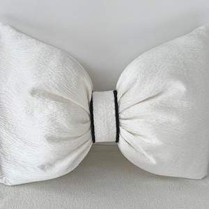 Könnte beinhalten: Ein großes, weißes Zierkissen in Form einer Schleife. Das Kissen hat eine strukturierte Oberfläche und ist in der Mitte mit einem schwarzen Band gebunden. Dieses Schleifenkissen ist ein dekoratives Element für ein Bett oder Sofa.
