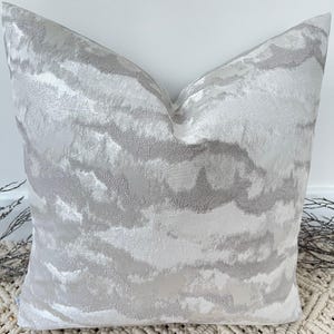 Op de afbeelding: Een decoratief vierkant kussen met een textuur, abstract ontwerp in grijstinten en wit. Het kussen heeft een zachte, pluche uitstraling en voegt een vleugje elegantie toe aan de woondecoratie. Het patroon lijkt op vloeiende wolken of penseelstreken.