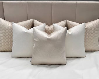 May include: A set of six decorative throw pillows in a neutral color palette. The pillows are arranged in a row on a bed with a light beige headboard. The pillows are made of a soft, textured fabric and have a subtle sheen.