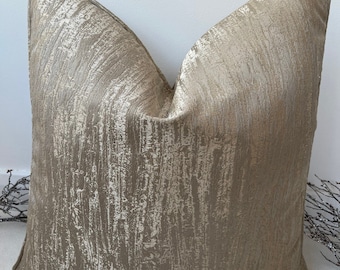 May include: A gold and beige throw pillow with a textured, brushed fabric. The pillow has a square shape and a subtle sheen.