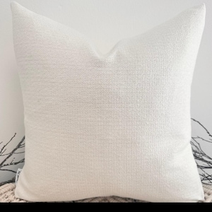 May include: A white, square cushion with a textured fabric. The cushion is sitting on a woven rug.