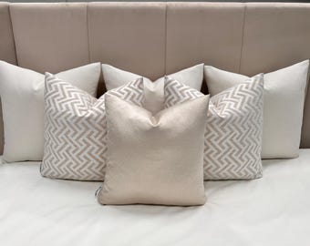 May include: A set of five throw pillows in a neutral color palette. The pillows are arranged on a bed with a light beige headboard. Two of the pillows have a geometric pattern in shades of beige and white. The other three pillows are solid beige.