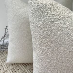 Op de afbeelding: Twee off-white kussens met een textuur in de stof. De kussens liggen op een geweven vloerkleed.