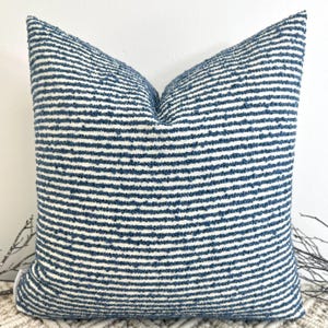 May include: A square cushion with a textured, horizontal striped pattern in navy blue and white. The cushion has a soft appearance, suitable for home decor. The dimensions are not visible.
