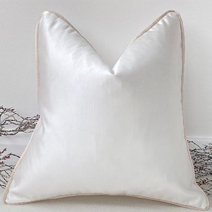 May include: A white cushion with a cream coloured border. The cushion is made of a soft, silky fabric.