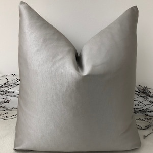 Könnte beinhalten: Ein silbergraues Kissen mit einer dezenten, strukturierten Oberfläche. Das Kissen ist quadratisch und hat ein schlichtes Design.