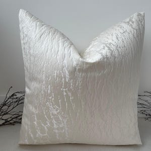 May include: A square, off-white decorative pillow with a textured, crinkled fabric. The pillow has a soft, neutral color and a subtle sheen, adding a touch of elegance to any room. The pillow is a home decor item.
