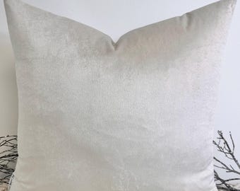 Könnte beinhalten: Ein quadratisches, hellbeige Samtkissen. Das Kissen hat ein weiches, strukturiertes Aussehen und einen subtilen Glanz. Das Kissen liegt auf einer weißen Makramee-Oberfläche, mit einer weißen Wand im Hintergrund.