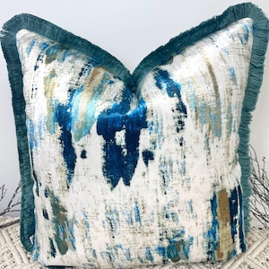Puede incluir: Cojín decorativo cuadrado con un diseño abstracto en tonos azules y dorados sobre fondo blanco. Cuenta con un ribete de flecos turquesa en los bordes. Este cojín es un elemento de decoración para el hogar.