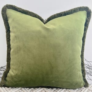 May include: A square, olive green velvet pillow with a fringe trim. The pillow has a soft, plush appearance and a darker green fringe around the edges. The pillow is a decorative home accent.