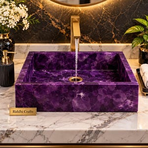 May include: A square bathroom sink crafted from deep purple amethyst stone with a polished finish. A gold-toned faucet is centered above the basin, with water flowing into the drain. A small gold plaque labeled Riddhi Crafts sits nearby.